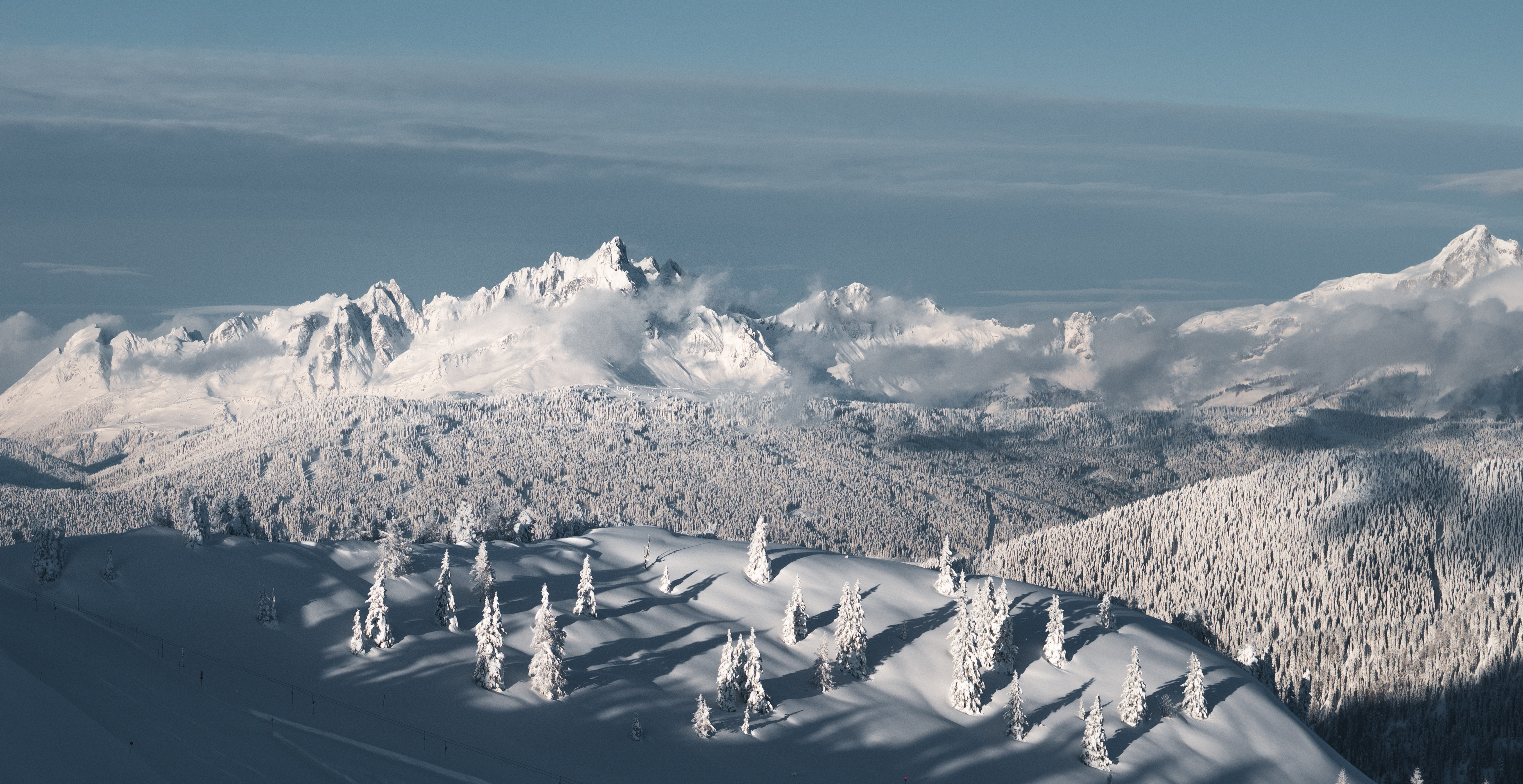 Vue panoramique de montagnes enneigées et de forêts de sapins gelés sous un ciel bleu clair, avec des ombres longues.
