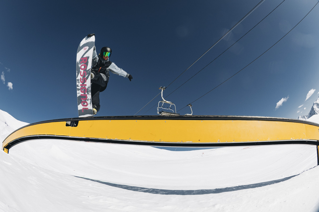 Snowboarder exécutant un grab en l'air sur une rampe jaune, entouré de neige immaculée et sous un ciel azur ensoleillé.