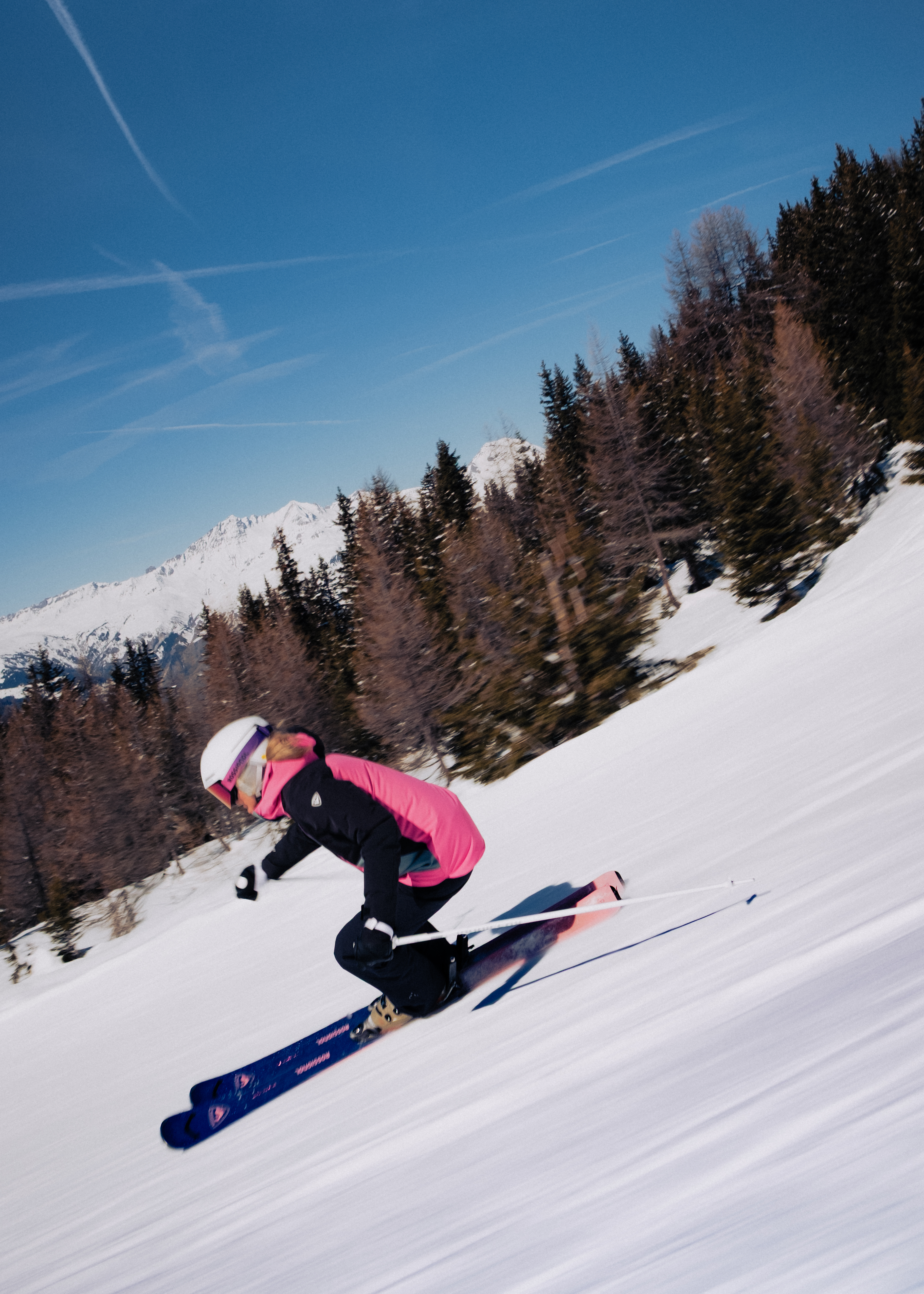 Une skieuse en veste rose et noire dévale une pente enneigée. Des sapins, des montagnes enneigées et un ciel bleu se trouvent en arrière-plan.