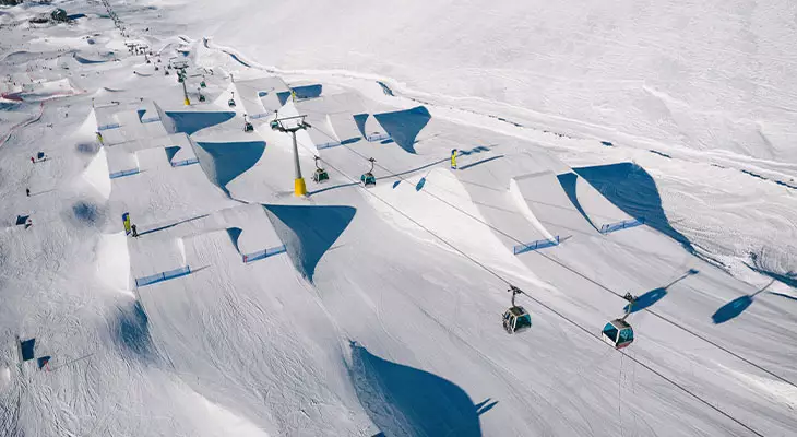 Vue aérienne d'un snowpark alpin enneigé avec de multiples tremplins, half-pipes et télécabines. Des skieurs sont visibles.