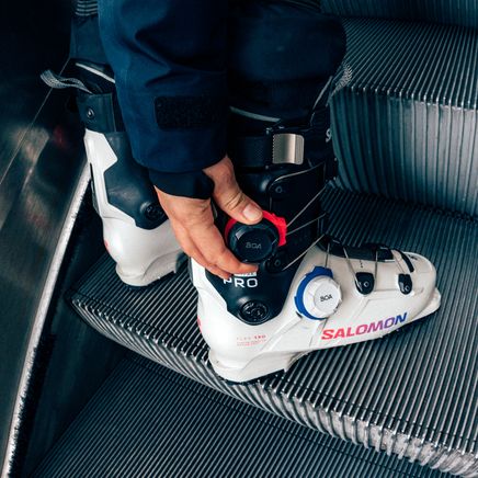 Une personne en chaussures de ski Salomon blanches et noires ajuste sa botte sur un escalator.