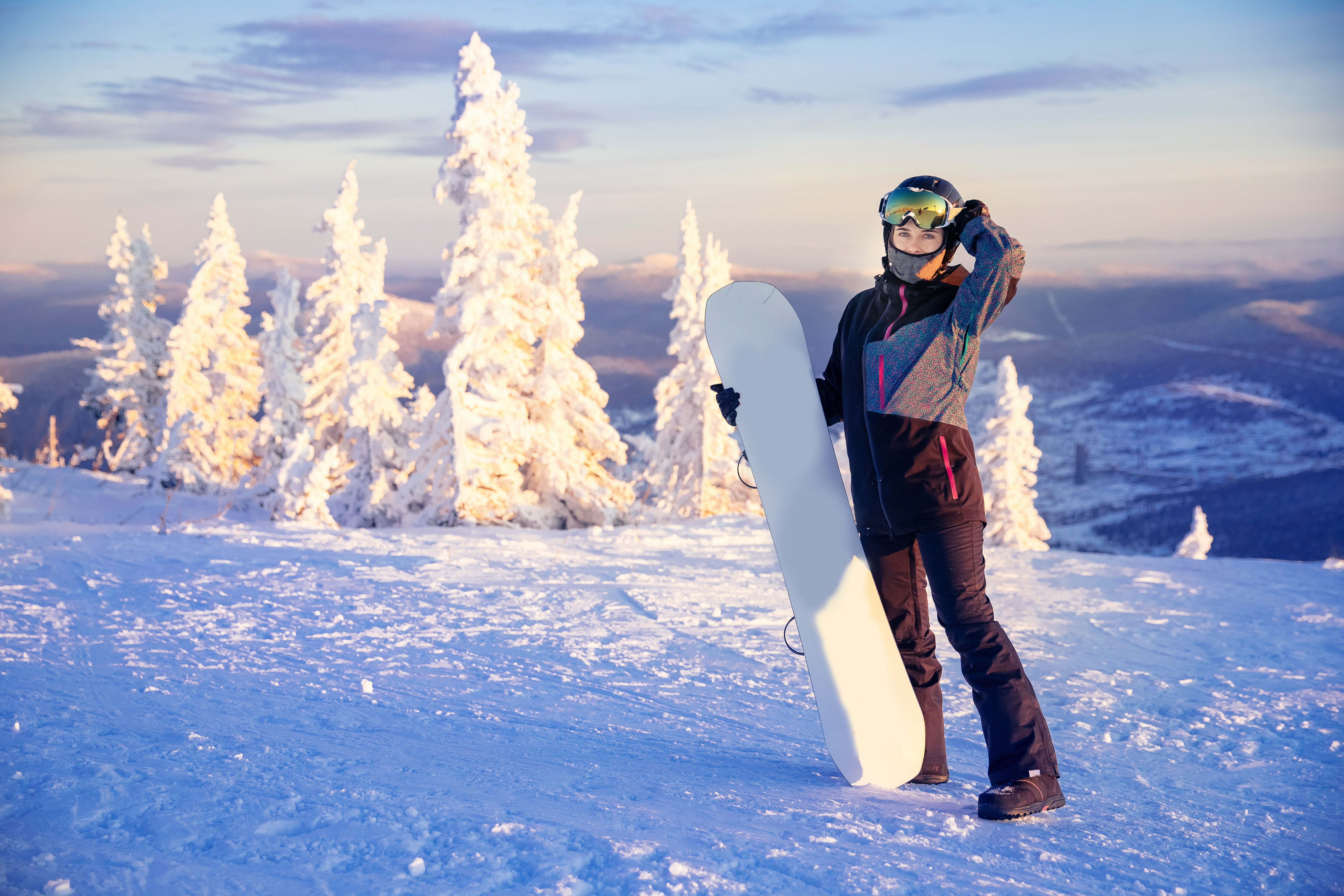 Femme snowboardeuse en tenue d'hiver sur une montagne enneigée, avec un snowboard blanc et des sapins givrés sous un ciel de coucher de soleil.