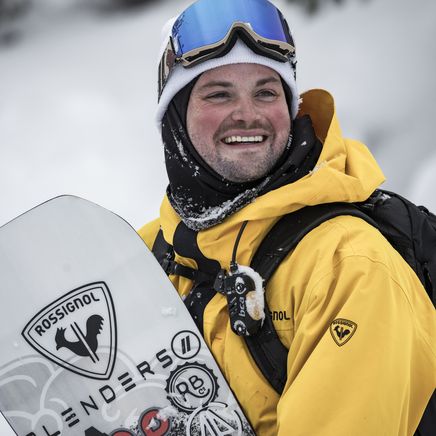 Homme snowboarder souriant, veste jaune, bonnet blanc, masque bleu, tenant une planche blanche sur fond enneigé.