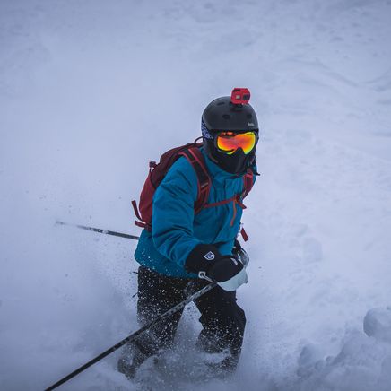 Skieur en veste bleue et masque orange, dévalant une pente enneigée, soulevant de la poudreuse. Une GoPro est sur le casque.