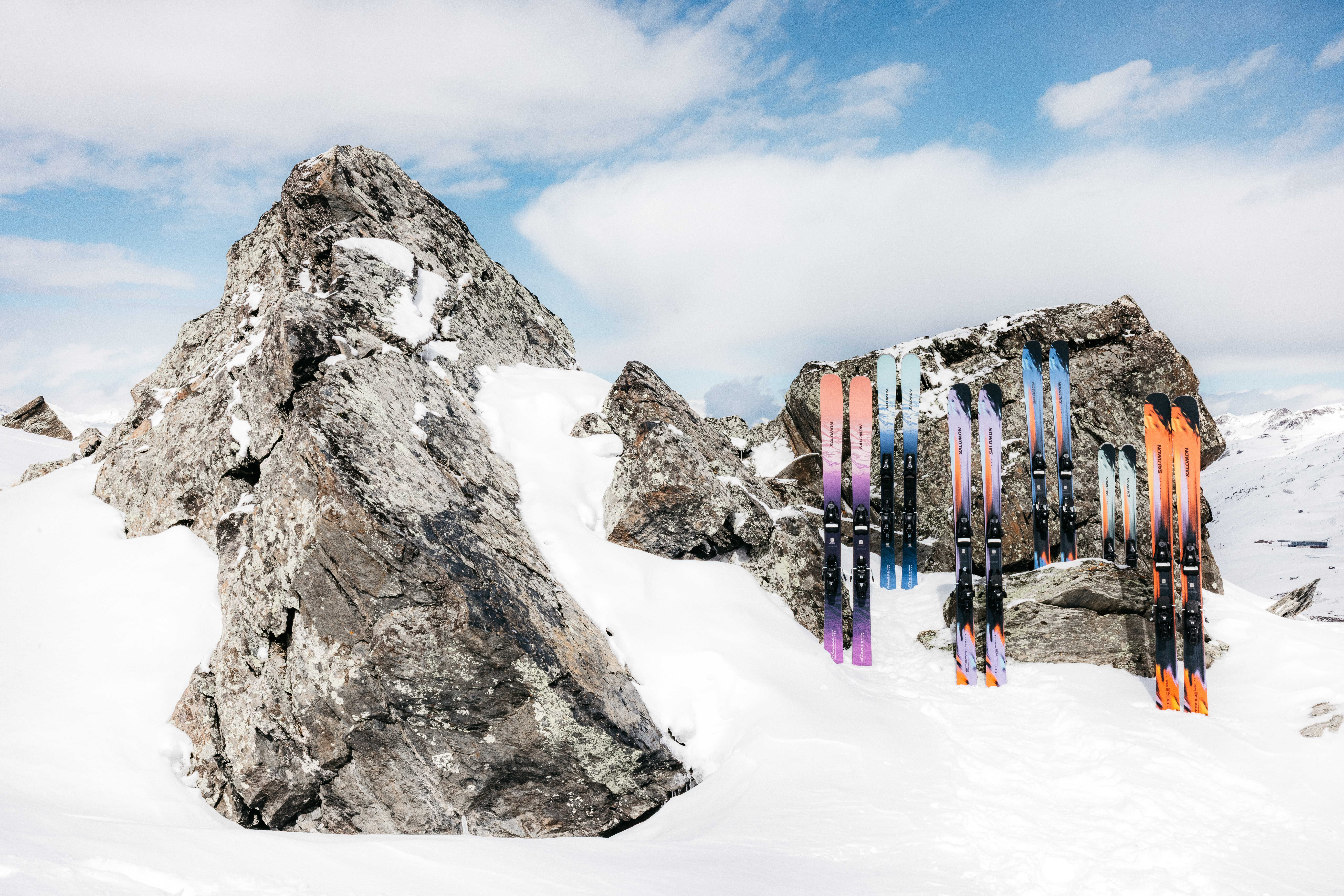 Plusieurs paires de skis colorés plantés dans la neige, appuyés contre des rochers dans un paysage montagnard enneigé sous un ciel bleu nuageux.