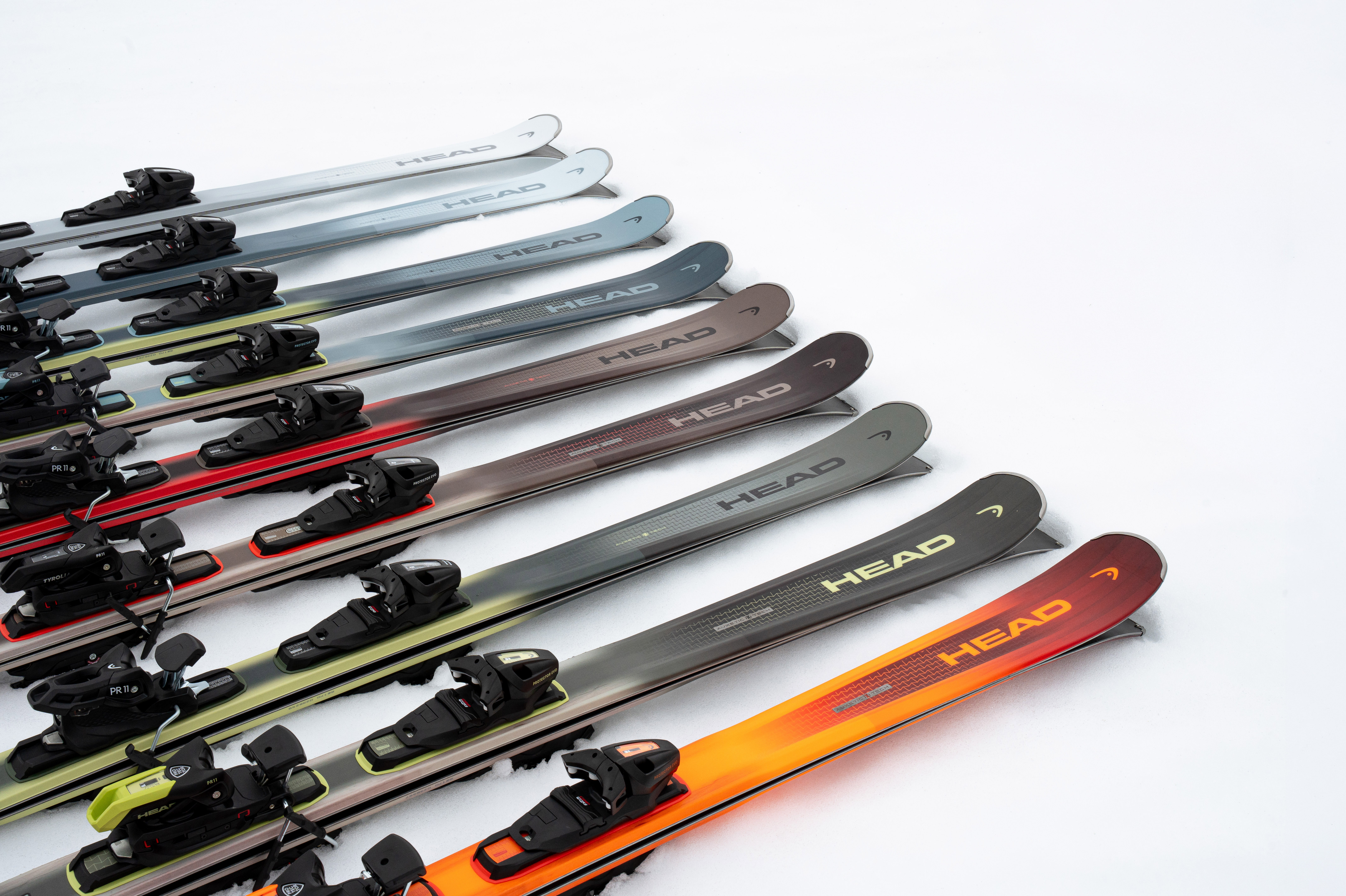 Plusieurs paires de skis Head de couleurs variées (bleu, gris, marron, rouge) alignées en diagonale sur la neige.