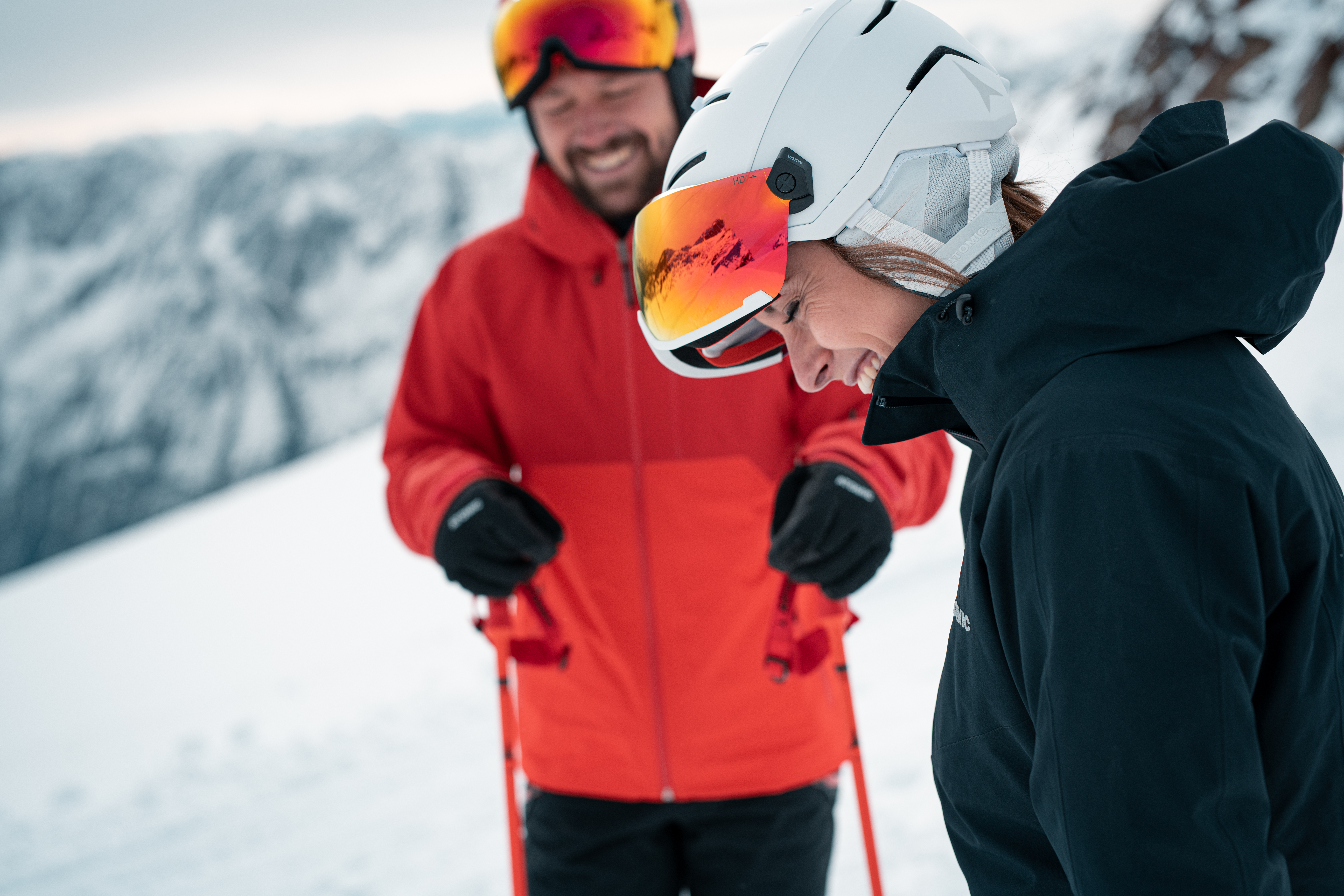 Deux skieurs souriants sur une piste enneigée. La femme en casque blanc et veste noire regarde vers le bas en riant, l'homme en rouge derrière.
