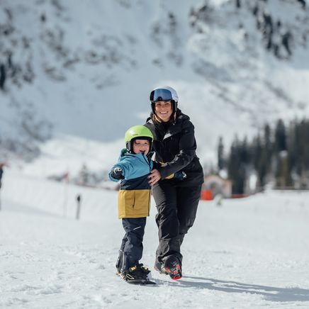 Femme souriante en tenue de ski aidant un enfant à skier sur une piste enneigée, montagnes et forêts en arrière-plan.