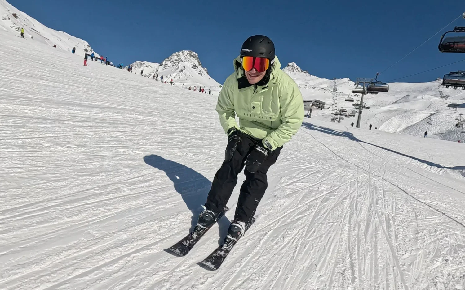 Skieur souriant en tenue verte dévalant une piste de ski enneigée, sous un ciel bleu éclatant, avec des montagnes et télésièges au loin.