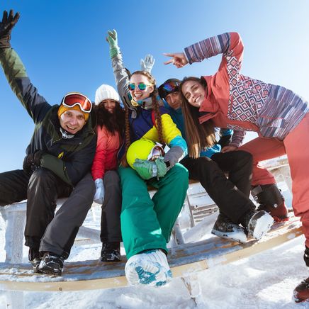 Groupe de six amis en tenues de ski colorées, posant gaiement sur un banc enneigé par une journée ensoleillée en montagne.
