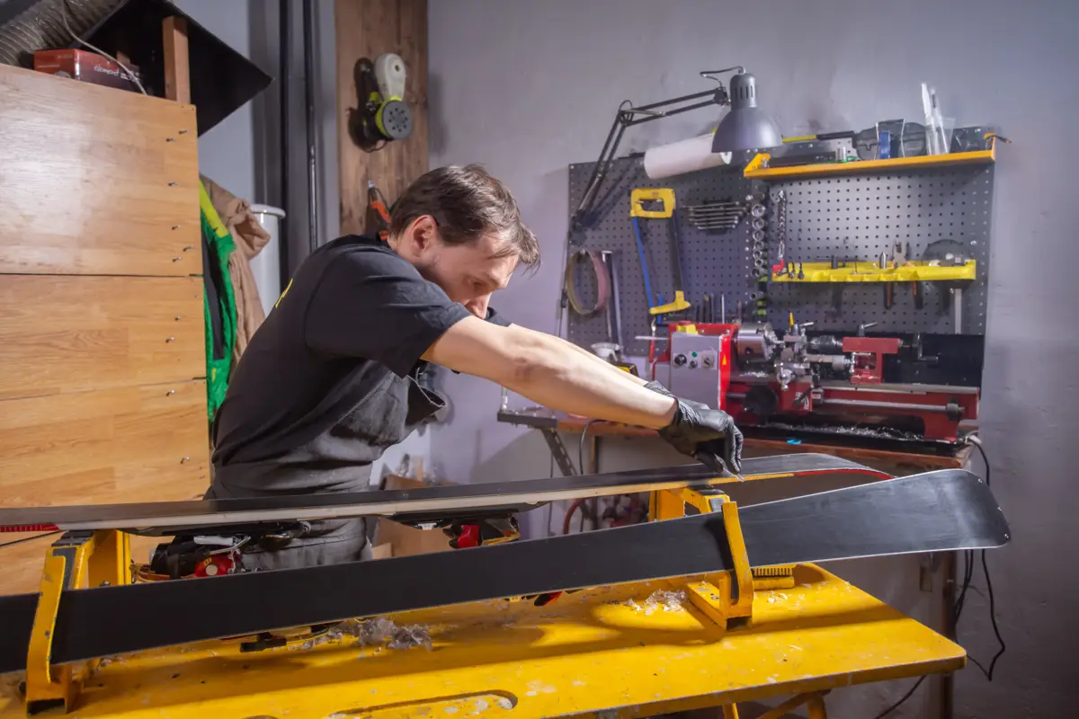 Un homme cire des skis sur un support jaune dans un atelier bien équipé.
