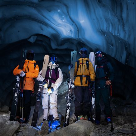 Quatre skieurs équipés posent avec leurs skis dans une grotte de glace bleue. Les murs sont de glace ondulée et brillante.