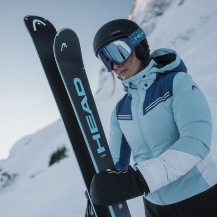 Femme skieuse en tenue bleue et noire, avec casque et masques, tenant des skis Head noirs et bleus sur une montagne enneigée.