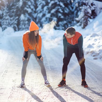 Homme et femme en tenue de sport font une pause, leur souffle visible, sur une route enneigée en plein soleil.