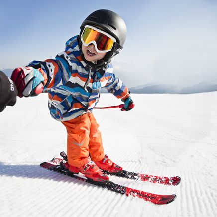 Un jeune enfant skie sur une piste enneigée, tenant la main d'un adulte. Il porte un casque, des lunettes et une veste colorée.