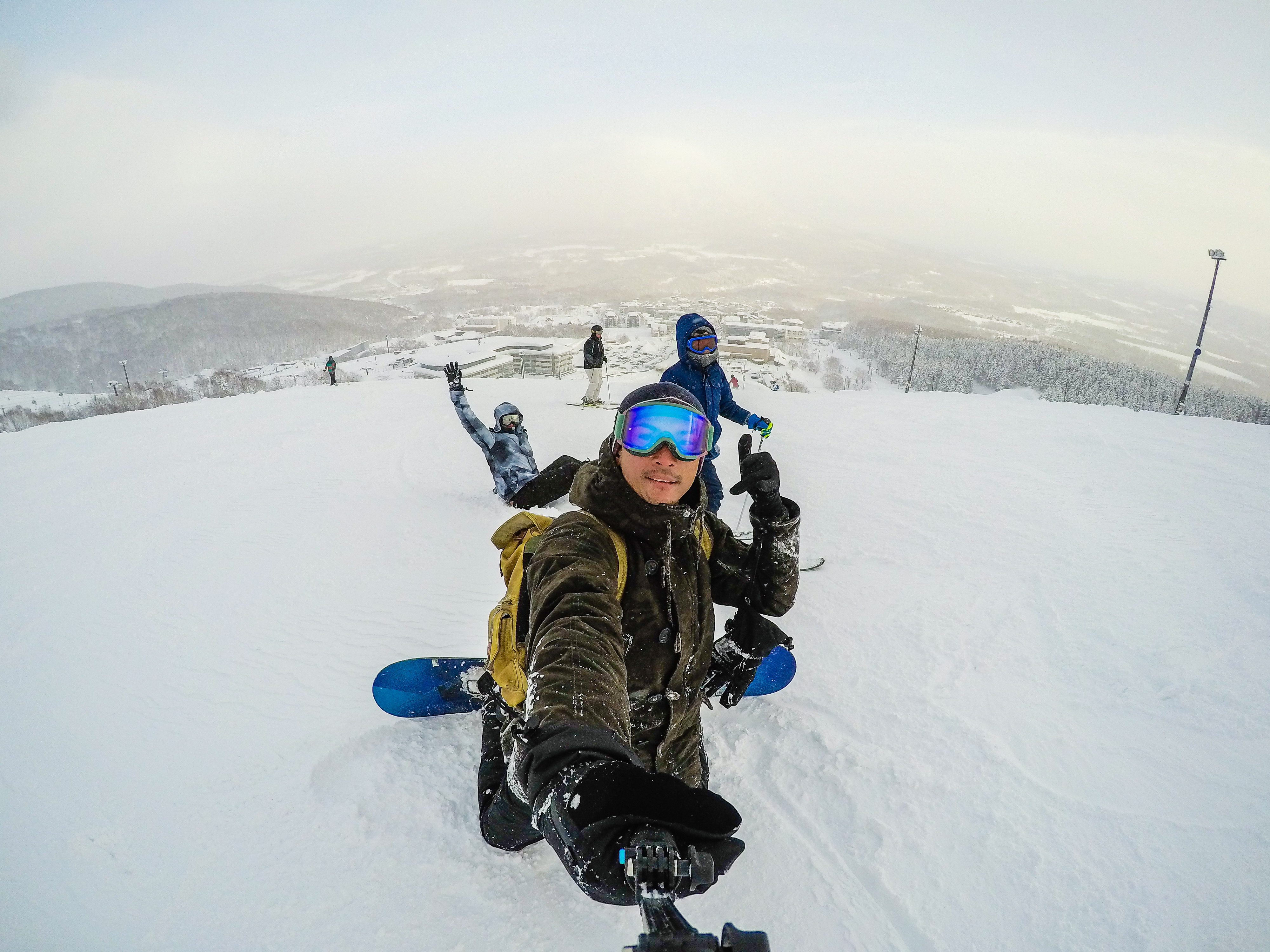 Un snowboarder prend un selfie avec des amis sur une piste enneigée, faisant un pouce levé. Vue sur un vaste paysage montagneux hivernal.